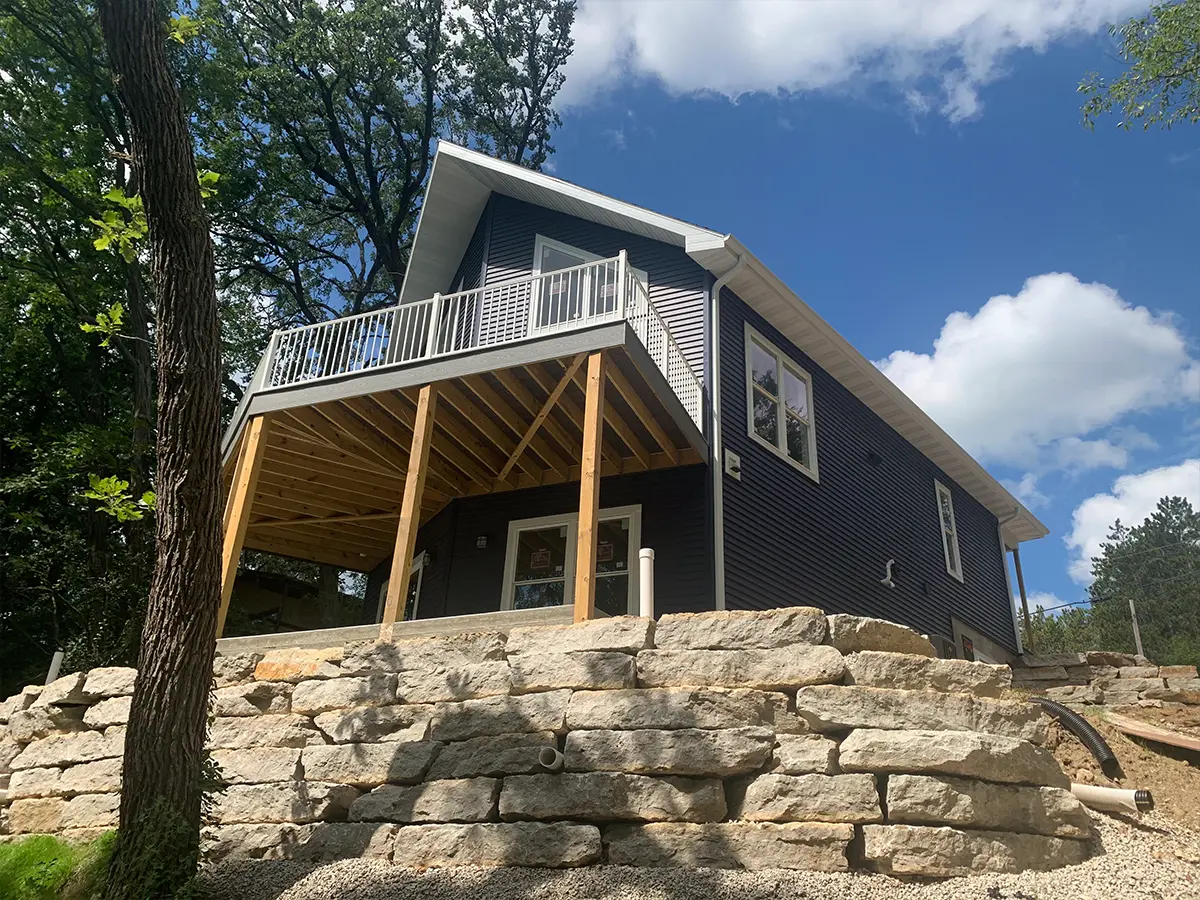 A retaining wall in a backyard with an elevated deck attached to a big home with dark blue siding